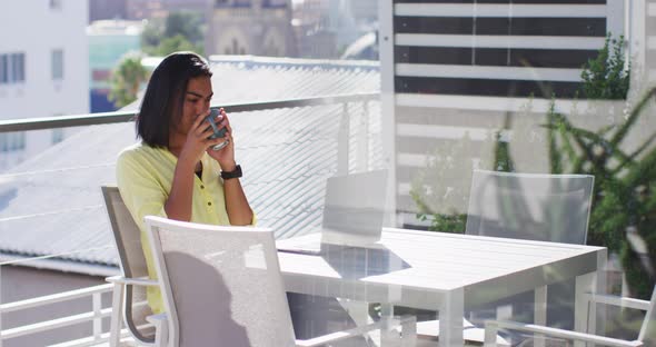 Mixed race gender fluid person drinking cup of coffee and using laptop on roof terrace alt