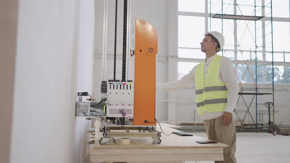 An Engineer Man in a Helmet Adjusts the Work of the Printer Printing on the Wall of the Building alt