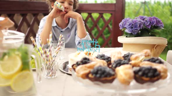 Sad Boy on His Birthday. A Lonely Teenager in a Birthday Hat Sits at a Table with a Treat and Cake alt