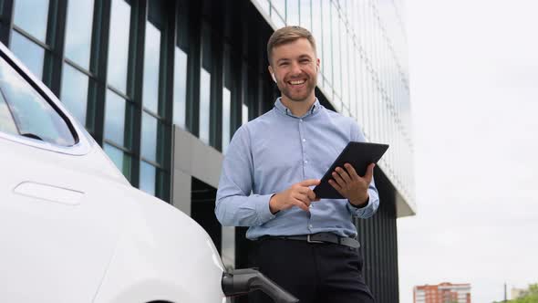 Young European Businessman with Wireless Headphones and Tablet Near Electric Car Charging the alt