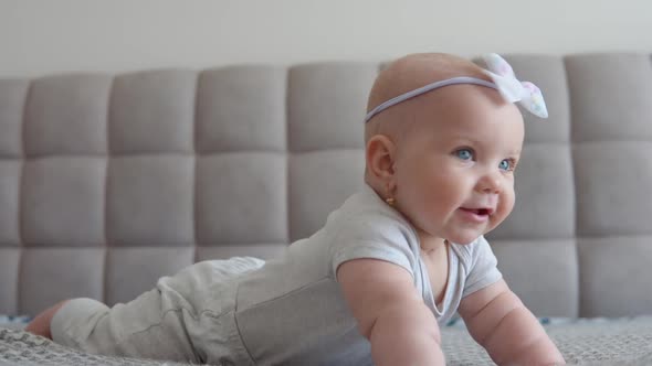 Baby Girl in Gray Clothes with a Cute Bow on His Head Lying on the Bed Tummy Down and Looking at the alt