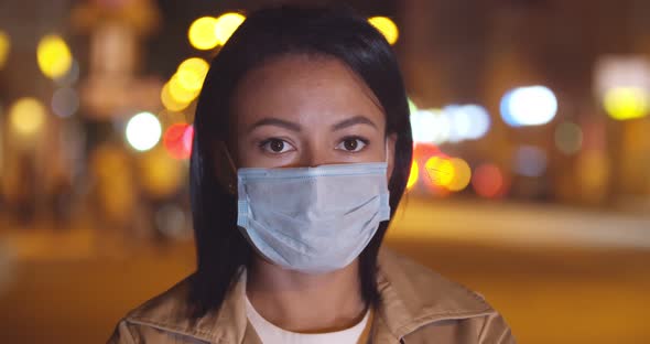 Bokeh Shot of Young Afro Woman in Safety Mask Looking at Camera Standing Outdoors alt