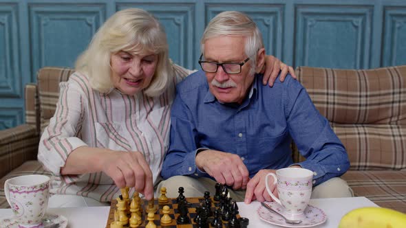 Smiling Senior Couple Grandfather Grandmother Resting on Sofa Drinking Coffee Playing Chess at Home alt
