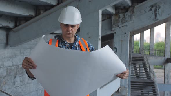 A Male Builder Unfolds and Examines the Drawing of a Building on the Construction Site