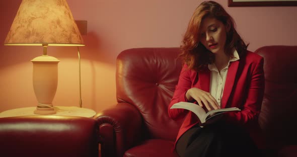 Beautiful woman with red jacket reads the book sitting on the sofa at home alt