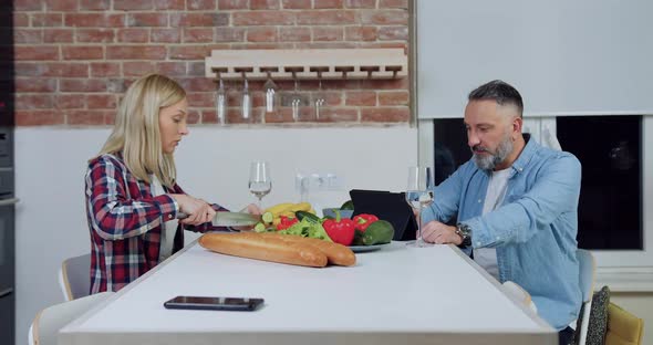 Man Working on Tablet PC and Eating Pepper Slice while His Smiling Adorable Blond Wife alt