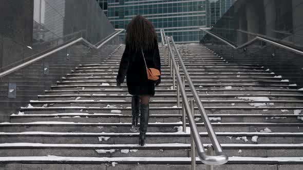 A Curlyhaired Woman in a Fur Coat Climbs the Stairs in the City Enjoying a Beautiful Day alt
