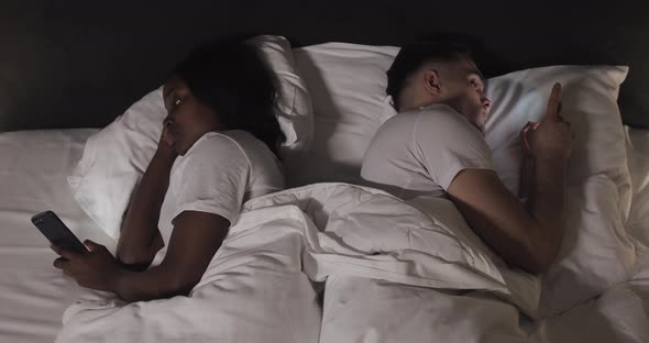 Married Young Multi-ethnic Couple Lying in the Bed Back To Back at Night and Typing or Scrolling  alt