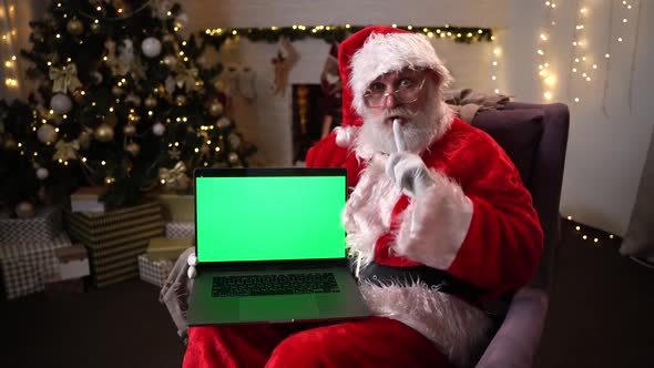 Santa Claus in Glasses Sitting in His Rocker Near Christmas Tree. Holds a Laptop with Green Screen