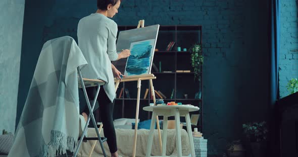 Young Woman Paints a Landscape on Canvas in a Bright Workshop alt