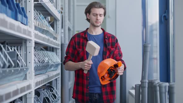 Positive Handsome Man Hitting Hard Hat with Hammer and Looking at Camera Standing in Hardware Store alt