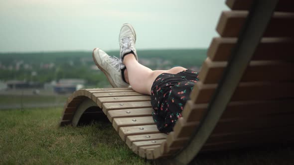 A Young Girl in a Dress and Sneakers Lies on a Deck Chair on the Grass alt