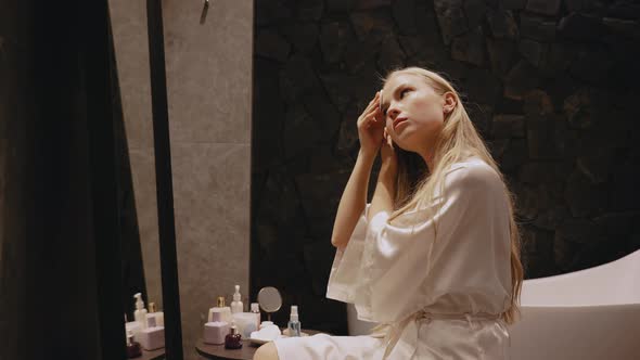Young Blonde in a Bathroom Sitting in Front of a Mirror Wipes Her Neck and Face alt