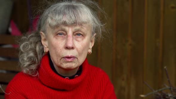 An Old Gray-haired Woman Smokes a Cigarette Sitting in Front of Her Dilapidated Home alt