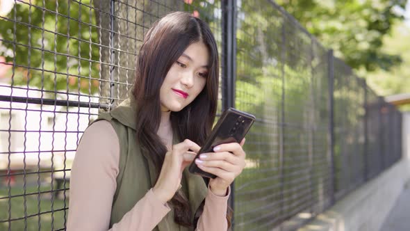 Young Asian Woman Works Smartphone Smile As She Leans Against Tall Fence alt