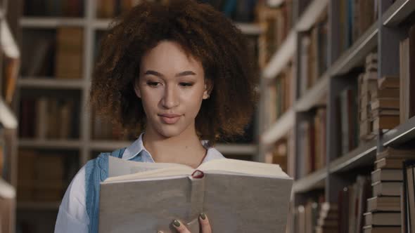 Young African American Girl Student Stands in Public University Library Interested in Reads Book alt