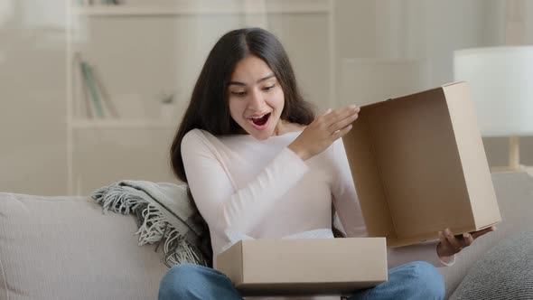 Surprised Arab Latin Woman Satisfied Customer Shopaholic Sits on Couch at Home Open Delivery Parcel alt