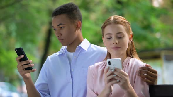 Hugging Multiethnic Teen Couple Sitting on Bench in Park, Chatting Smartphone alt