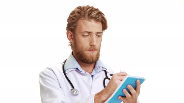 Young Handsome Caucasian Male Doctor Making Notes in Blue Notebook and Looking at Camera on White