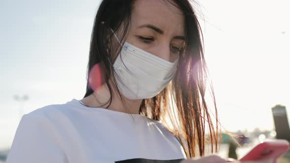 Young Woman in a Medical Mask Use Smartphone for Protection From the Epidemic on the Street in the alt