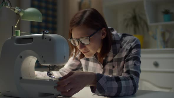 Female seamstress in glasses sewing sitting in living room at home alt