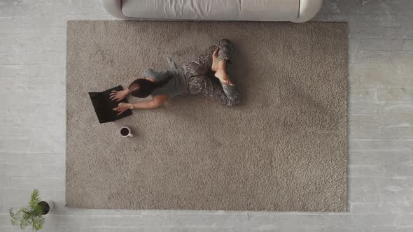 A Young Woman Is Lying on the Carpet Drinking Coffee and Using a Laptop