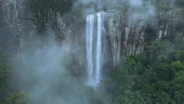Cinematic view looking through rising mist revealing a majestic waterfall spilling over a lush rainf alt
