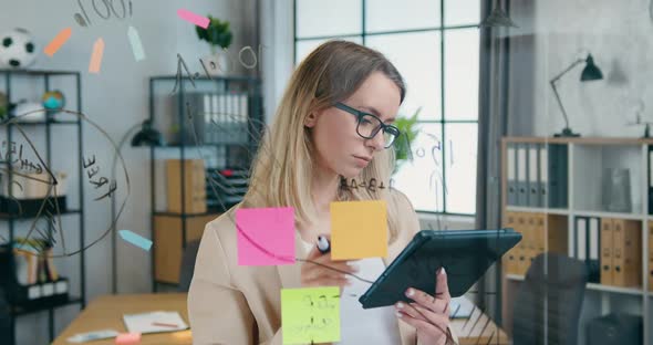 Woman in Glasses which Standing Near Glass Board and Writing Datas from Tablet PC alt