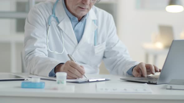 Male Doctor Taking Notes and Working on Laptop in Medical Office alt