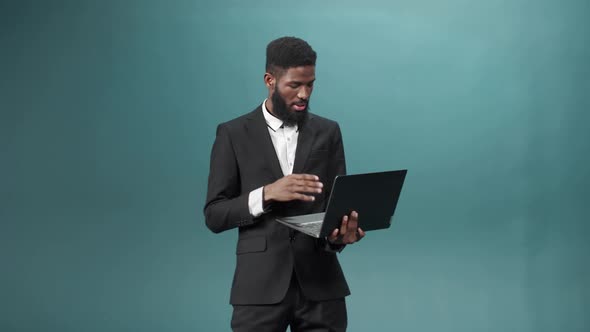 An Attractive African Bearded Man is Standing in a Tuxedo Keeping a Laptop in His Hands and Talking