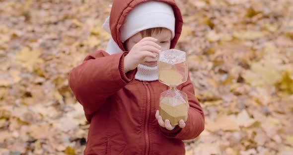 Little a Caucasian Girl is Playing with a Glass Hourglass in the Autumn Forest