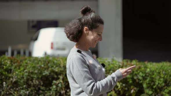 Side View of Middle Aged Woman Walking on Street with Phone. alt