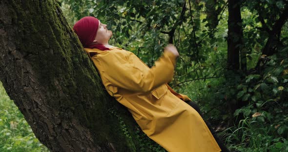 Hiker in Bandana and Yellow Raincoat is Resting in Forest Leaning Against Tree