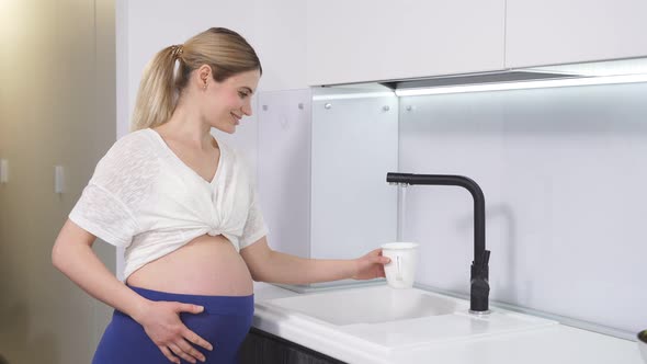 Side View of a Pregnant Woman Standing at the Tap and Pouring Water Into a Drinking Glass After a