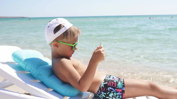 Child Plays on a Tablet While Sitting on the Beach on a Sunny Summer Day