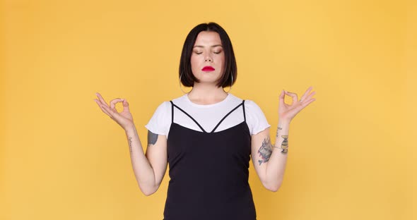 Young hipster woman with tattoos meditates and relaxes on a yellow background. alt