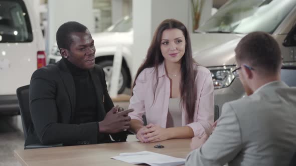 Happy Young Multiracial Couple Sitting in Car Dealership and Talking with Salesman alt