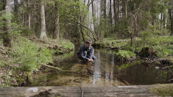 Man Sits in Overgrown Stream in Forest Picks Up Cool Water and Washes His Face alt