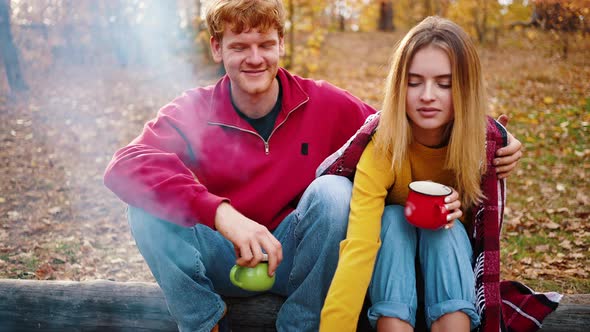 Young Man Hugging Girlfriend Who Holding Cup of Tea While Sitting on Log in Autumn Wood alt