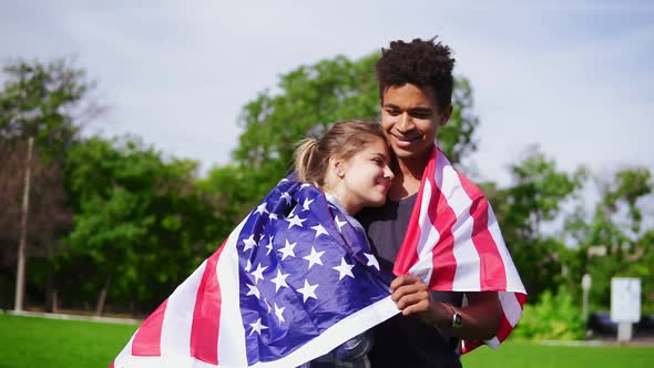 Attractive Multiethnic Couple Embracing Each Other Holding American Flag on the Back Standing in the alt