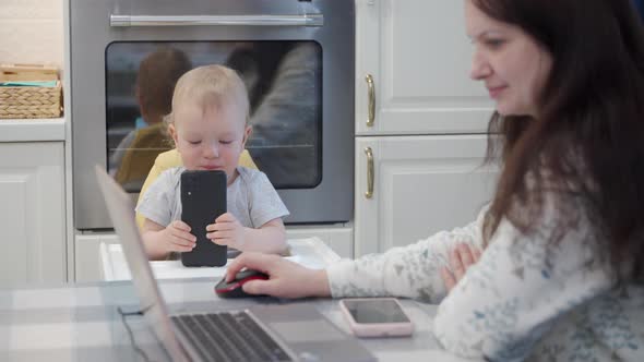 Mother Works on Laptop While Her Toddler Playing with Smartphone One Year Old Baby Boy Sitting in alt