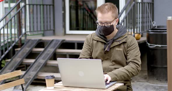 A man in a mask works on a laptop while sitting in a cafe on the street alt
