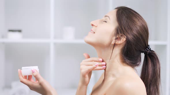 Smiling Woman Enjoying Applying Moisturizing Cream on Neck at White Bathroom Light Background alt