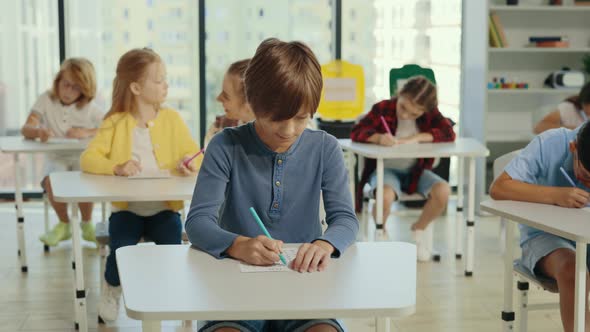 Male Pupil Sits at a First Desk in the Classroom and Fills Out Tests Thinking and Writing Down alt