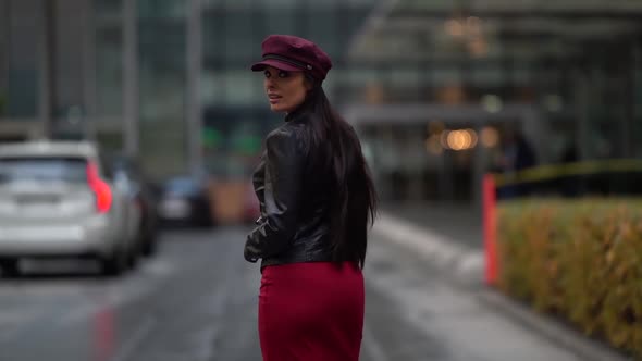 a Brunette with Long Hair in a Black Leather Jacket a Red Skirt and a Burgundy Cap Walks Against the alt