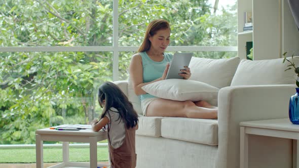 Young mother chatting with tablet and daughter drawing on paper