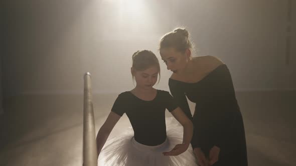Medium Shot of Slim Woman Explaining Third Ballet Position Talking with Girl in Dance Studio Backlit alt
