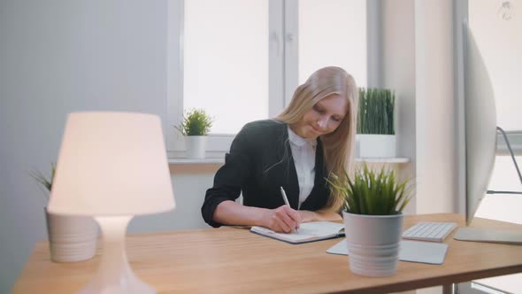 Beautiful Young Blond Female Suit Sitting at Desk Writing with Pen in Notepad, Turning Page and alt
