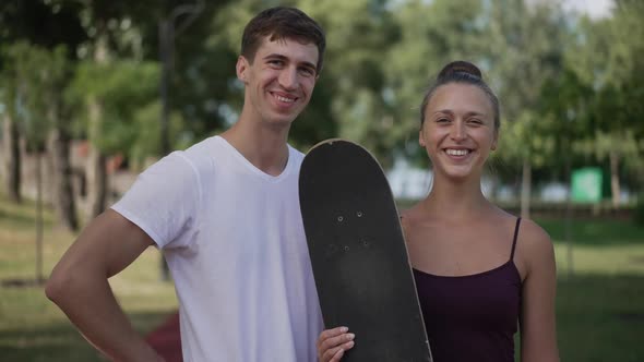 Happy Loving Young Couple with Skateboard Looking at Each Other Turning to Camera Gesturing Thumbs alt