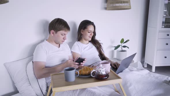 Woman with Laptop Sits Near Husband Using Smartphone on Bed